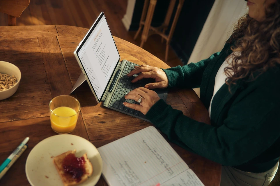 Mujer escribiendo en laptop en mesa de madera con desayuno.